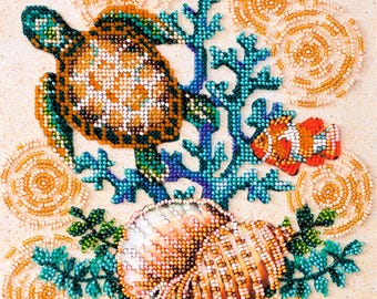 Könnte beinhalten: Ein Perlenstickerei-Kunstwerk mit einer Meeresschildkröte, einem Clownfisch und einer großen Muschel inmitten blauer Korallenzweige. Das Design ist mit kreisförmigen Mustern aus cremefarbenen Perlen auf hellem Stoffgrund verziert.