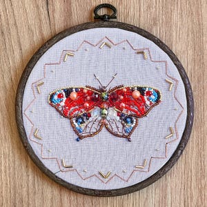 May include: A red, blue and gold beaded butterfly embroidery on a white fabric background in a round wooden embroidery hoop.