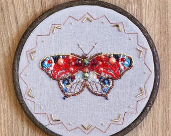 Könnte beinhalten: Eine rote, blaue und goldene Perlen-Schmetterlings-Stickerei auf einem weißen Stoffhintergrund in einem runden Holz-Stickrahmen.