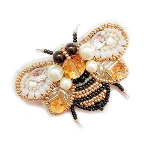 May include: A beaded bee brooch featuring black, gold, white and clear beads. The bee has a detailed body with a black and gold striped abdomen and clear and gold bead wings.