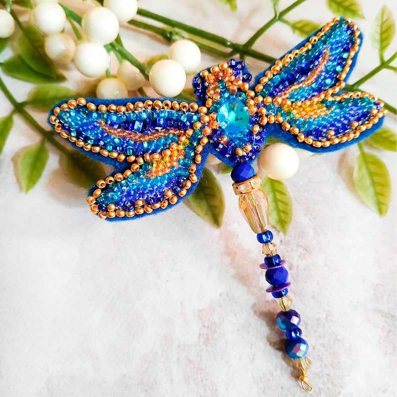 Pode incluir: Um broche de lib&eacute;lula azul e dourado com contas, com um detalhe de cristal e uma corrente pendente de contas azuis e transparentes.