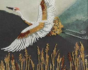 May include: A beaded artwork depicting a crane in flight. The bird is white, brown, and gold, with outstretched wings. The background features a dark landscape with tall, beaded grasses in the foreground.