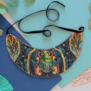 May include: A beaded bib necklace featuring a scarab beetle design. The necklace has a black velvet cord and is adorned with blue, green, gold, and orange beads. The design is reminiscent of ancient Egyptian jewelry.