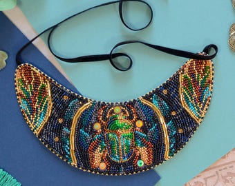 May include: A beaded bib necklace featuring a scarab beetle design. The necklace has a black velvet cord and is adorned with blue, green, gold, and orange beads. The design is reminiscent of ancient Egyptian jewelry.