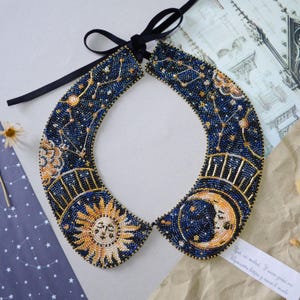 Könnte beinhalten: Ein mit Perlen besetztes Kragenhalsband mit einem Himmelsdesign. Der marineblaue Kragen zeigt goldene, weiße und gelbe Perlenarbeiten, die Sternbilder, eine Sonne und einen Mond darstellen. Ein schwarzes Band ist zum Verschließen befestigt. Ein einzigartiges Modeaccessoire.