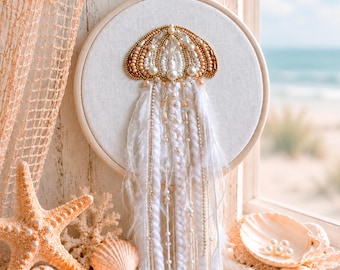 May include: A circular embroidery hoop containing a beaded jellyfish design on white fabric. The jellyfish features gold, white, and cream beads with long, dangling white yarn, feathers, and beaded strands hanging from the bottom of the hoop.