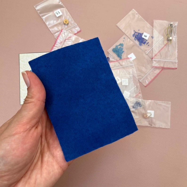 Pode incluir: Uma almofada de feltro azul para artesanato e fabrica&ccedil;&atilde;o de joias. A almofada est&aacute; sobre uma superf&iacute;cie rosa com pequenas bolsas de contas e outros materiais de artesanato.