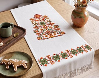 May include: A white rectangular table runner featuring a central cross-stitch tree motif and a geometric border pattern in shades of green, orange, and brown. The runner is finished with a fringed edge and is displayed on a wooden surface.