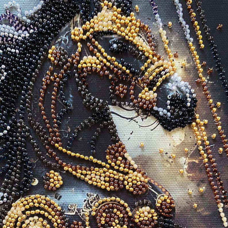 Puede incluir: Primer plano de una obra de arte con cuentas que representa la cabeza de un caballo. El caballo est&aacute; adornado con intrincados abalorios en tonos negros, marrones y dorados. El fondo es un lienzo oscuro y texturizado, que realza el dise&ntilde;o de cuentas.