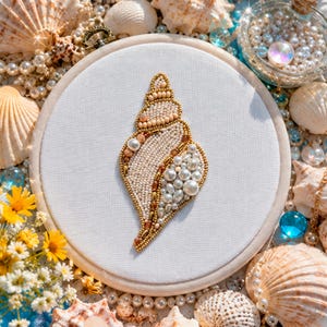 May include: An embroidered seashell design on white fabric, set in a wooden hoop. The shell is decorated with cream, gold, and pearl beads. The background features seashells, pearls, and small yellow flowers, evoking a seaside theme.