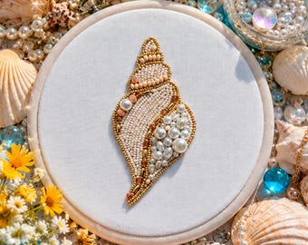Könnte beinhalten: Stickerei einer Muschel auf weißem Stoff, eingefasst in einem Holzreifen. Das Muscheldesign weist cremefarbene, goldene und Perlen auf. Der Hintergrund zeigt Muscheln, Perlen und kleine gelbe Blumen, die ein Küstenmotiv erzeugen.
