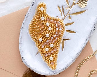 Könnte beinhalten: Eine goldene und weiße Perlenbrosche in Form eines Blattes, auf einer weißen Keramikplatte mit goldenen Akzenten.