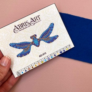 Pode incluir: Um close-up de um kit de pintura de diamantes com um design de lib&eacute;lula azul e dourado. O kit inclui uma tabela de cores com 16 cores diferentes e o texto "ABRISART www.abrisart.com AD-073".
