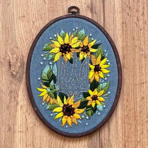 May include: Embroidered hoop art featuring a blue and yellow Ukrainian trident surrounded by yellow sunflowers on a blue background.