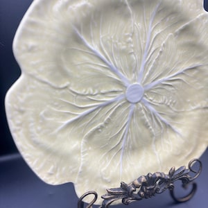 Peut inclure: Une assiette en céramique jaune pâle en forme de feuille de chou avec des nervures blanches en relief rayonnant depuis le centre. L'assiette est présentée sur un support en métal sombre avec des détails de volutes sur la base.