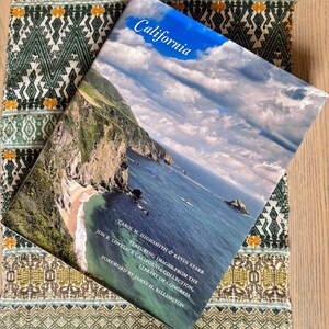 May include: A hardcover book titled California by Carol M. Highsmith and Kevin Starr, featuring a photograph of a rugged coastline with cliffs, a sandy beach, and the ocean under a blue sky with clouds. The cover includes text about the collection.