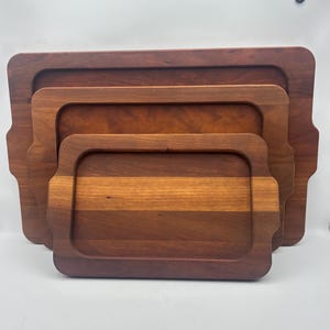 May include: A set of three rectangular wooden serving trays nested together. Each tray features a deep, recessed center, rounded corners, and integrated side handles. The wood displays a rich, warm brown grain with natural color variations.