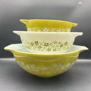 May include: Three vintage Pyrex nesting bowls in shades of green and white. The bowls feature a floral pattern in white. The bowls are stacked, with the largest bowl at the bottom and the smallest at the top. The bowls have handles.