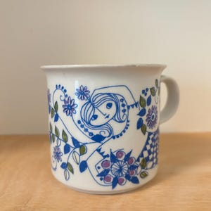 May include: A white ceramic mug featuring a blue and purple line art illustration of a person with long hair surrounded by stylized flowers, vines, and a checkered pattern. The mug has a simple handle on the right side and a smooth, rounded rim.