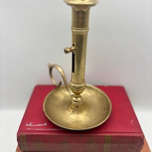 May include: A brass candlestick with a circular base and a handle, placed on two antique books with red and brown covers. The candlestick features a cylindrical stem and a small lever.