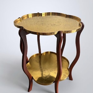 May include: A two-tier side table featuring a dark brown wooden frame with four curved legs. The table has two round, scalloped-edge brass trays with engraved floral patterns on the top and bottom surfaces, providing a tiered storage design.