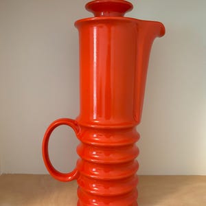 May include: A bright orange ceramic pitcher with a tall, cylindrical body and a ribbed, accordion-style base. The vessel features a curved handle on the side, a smooth upper section, a small pouring spout, and a matching circular lid on top.