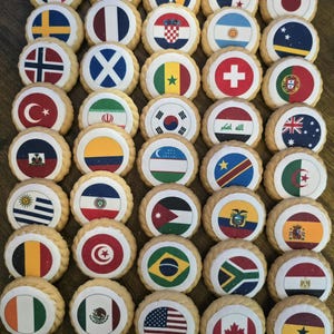 May include: A collection of round sugar cookies arranged in a grid, each featuring an edible image of a different national flag. The cookies have a scalloped edge and a smooth, white-frosted top surface displaying various colorful flag designs.