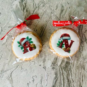 May include: Two round Christmas cookies in clear cellophane bags, tied with red ribbons. Each cookie has a white icing top with a letter and Santa hat design. The ribbon says "Merry Christmas".