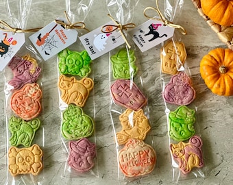 May include: Halloween-themed cookies in clear cellophane bags tied with gold twine. Each bag contains several cookies in the shape of pumpkins, cats, skulls, and tombstones. Tags with "Happy Halloween" and other Halloween phrases are attached.
