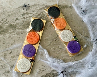 May include: Two packages of Halloween-themed cookies. Each package contains four round cookies with various designs, including a witch, a cat, a skull, and a pumpkin. The cookies are in clear cellophane bags with gold backing, and are decorated with black and orange icing.
