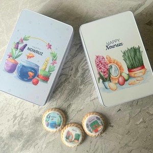 May include: Two rectangular metal boxes and three round cookies. The boxes have a white background with the words "Happy Nowruz" and illustrations of a fishbowl, flowers, and other festive elements. The cookies have similar designs.