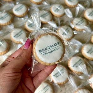 Op de afbeelding: Een hand houdt een doorzichtige cellofaan zak met een ronde koek met witte glazuur en de tekst "RACEMARK Manufaktur" in het groen. Andere koekjes met hetzelfde ontwerp zijn zichtbaar op de achtergrond.