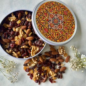 Op de afbeelding: Een ronde blik met een kleurrijk bloemmotief op de deksel, gevuld met een mix van gedroogde cranberries, rozijnen, amandelen en walnoten. De blik staat op een wit oppervlak met wat van de trailmix eromheen verspreid.
