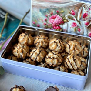 Puede incluir: Una lata rectangular azul claro llena de macarrón de coco con un chorrito de chocolate. La tapa de la lata tiene un diseño floral. Los macarrones dorados están dispuestos en el interior, ofreciendo una presentación tentadora.