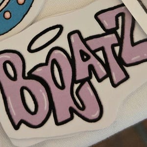 May include: A white sticker featuring the word "BRATZ" in a bubble letter font. The letters are light purple with a black outline and a white halo above the "B".