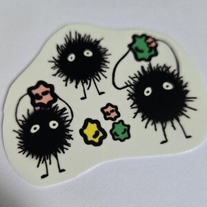 May include: A sticker featuring several black, fuzzy soot sprite characters with white eyes and small legs. The sprites are connected to colourful star-shaped objects. The sticker has a white background.