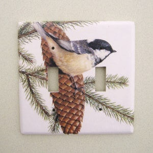 May include: A white ceramic light switch cover with a black-capped chickadee perched on a pine cone. The bird is brown, white, and black with a small, pointed beak. The pine cone is brown and has a green pine branch behind it.