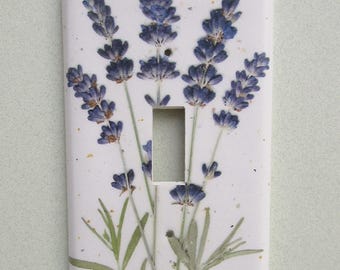 May include: A white light switch cover featuring a design of lavender sprigs. The lavender has purple flower heads and green stems and leaves. The switch cover has a rectangular shape with a single switch opening.