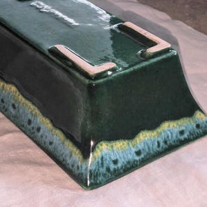 May include: A green ceramic planter with a light blue and yellow glaze. The planter has a rectangular shape with rounded edges and a raised lip around the top.