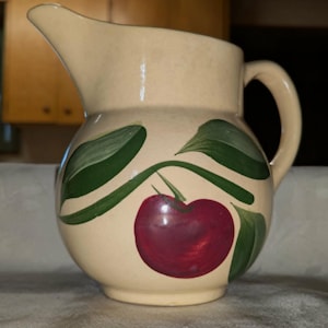May include: Cream-colored ceramic pitcher with a rounded body, spout, and handle. The pitcher is decorated with a large red cherry and green leaves. The pitcher is likely used for serving beverages.