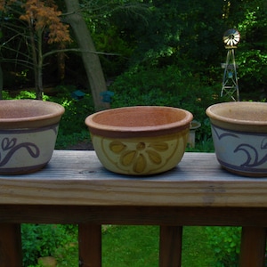 May include: Three ceramic plant pots with a rustic design. The pots are various shades of brown and beige, with decorative floral and swirl patterns in purple. The pots are sitting on a wooden deck.
