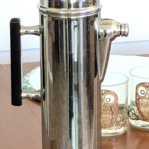 May include: A tall, cylindrical silver-toned metal cocktail shaker featuring a polished reflective finish, a black handle attached with silver hardware, a matching black knob on the lid, and a short, tapered pouring spout on the side.