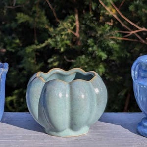 May include: Three ceramic planters in shades of blue and green sit on a white surface. The planters have unique shapes and textures, including a fluted design, a pumpkin-like form, and a decorative pattern. The background is blurred green foliage.