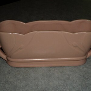 May include: A pale pink ceramic planter with a scalloped edge and two handles. The planter has a smooth, matte finish.