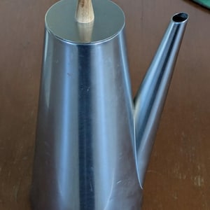 May include: A conical stainless steel oil cruet with a polished metallic finish, featuring a long, slender pouring spout and a flat circular lid topped with a small, light-colored wooden handle for easy lifting and controlled pouring of liquids.