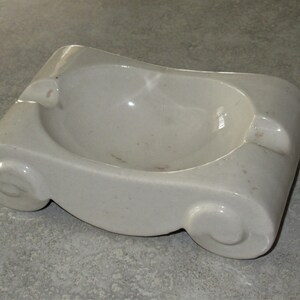 May include: A white ceramic ashtray with a scroll design. The ashtray has a round, indented center for holding cigarette butts.