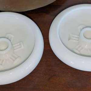May include: Two white ceramic candle holders with a circular design and a small handle. Each holder features a central well for a candle and a decorative embossed pattern. The holders are placed on a dark wooden surface.