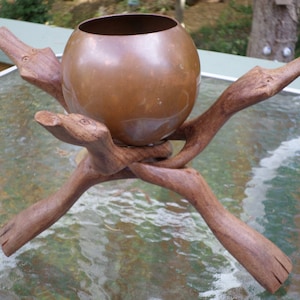 May include: A copper planter sits on a wooden stand with three legs. The stand is carved to resemble branches with bird heads at the ends.