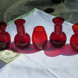 May include: Five red glass bud vases of different shapes and sizes. The vases are arranged in a row on a white tablecloth.