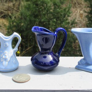 May include: Three small ceramic pitchers in shades of blue. The pitcher on the left is light blue with a floral design. The center pitcher is a deep cobalt blue. The pitcher on the right is a light blue vase with a flared top.